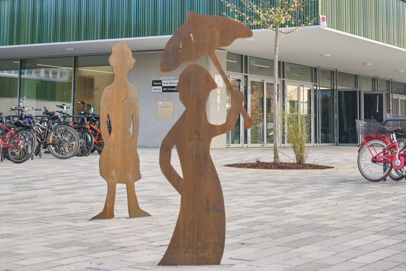 zwei Eisen-Skulpturen auf einem Hof vor einer Schule und Fahrrädern