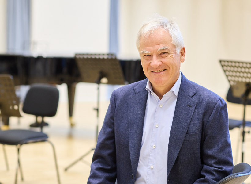 Altexander Pirlet sitzend in einem Klassenraum der Rheinischen Musikschule Köln vor mit Stühlen und Notenständern